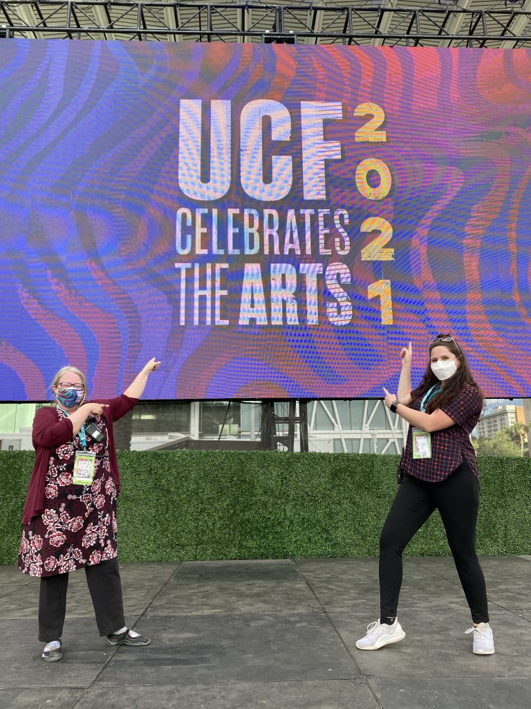 Claudia Lynch and Lauren Koval stand on a stage pointing to a screen that says "UCF Celebrates the Arts 2021"