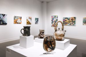 Four pieces of art, including a teapot and a mug, on display on a table in front of a wall with seven art pieces hung up at the Midnight Shift: Spring 2023 BFA Exhibition I.