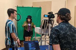A student stands in front of a green screen dressed as the Mad Hatter.